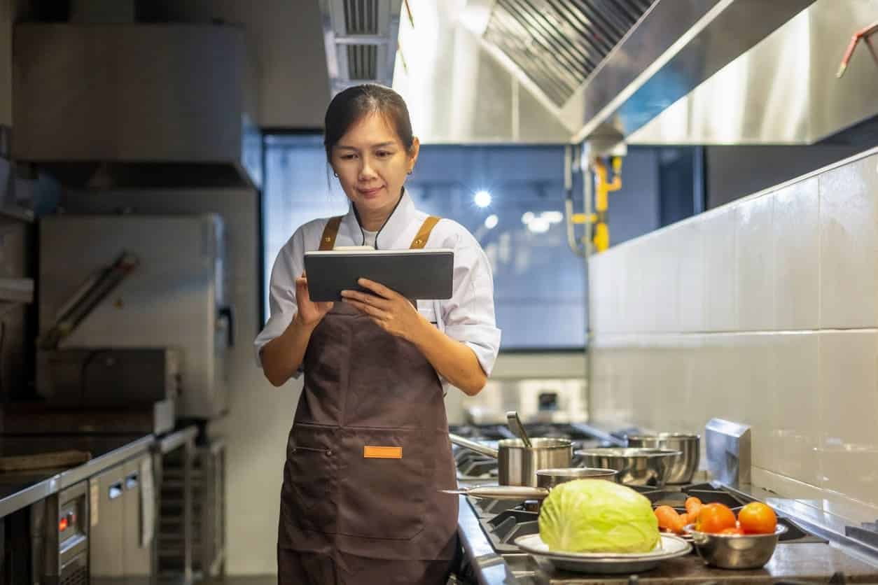 Cozinheira em cozinha industrial profissional, usando avental e segurando um tablet, ao lado de panelas no fogão e ingredientes como alface e laranjas sobre a bancada.
