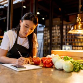 Mulher em cozinha de restaurante com verduras ao redor anotando em caderno para fazer o planejamento.