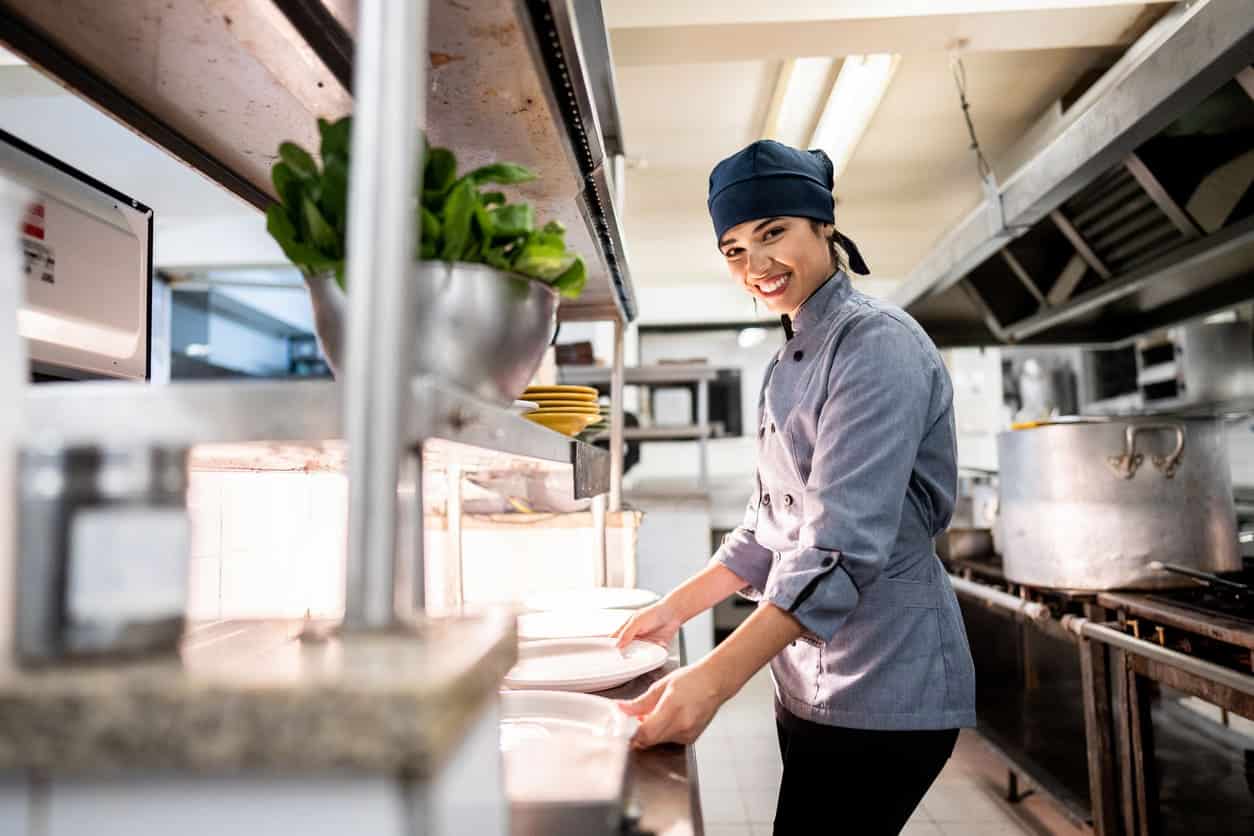 Chefe em pé na cozinha industrial segurando louças brancas e olhando para a foto.