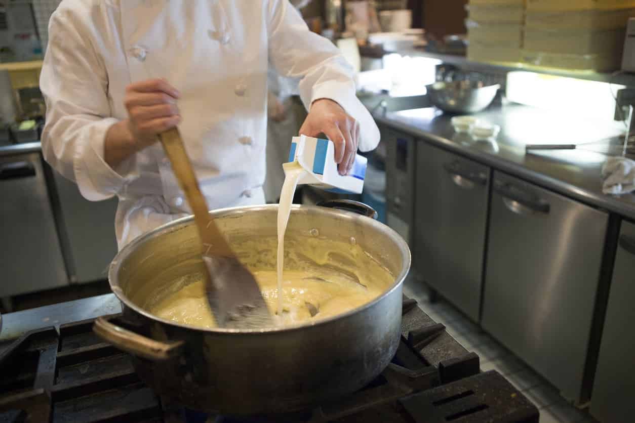 Chefe de cozinha preparando um molho bechamel, na cor branca, e adicionando leite em uma panela grande e prata.