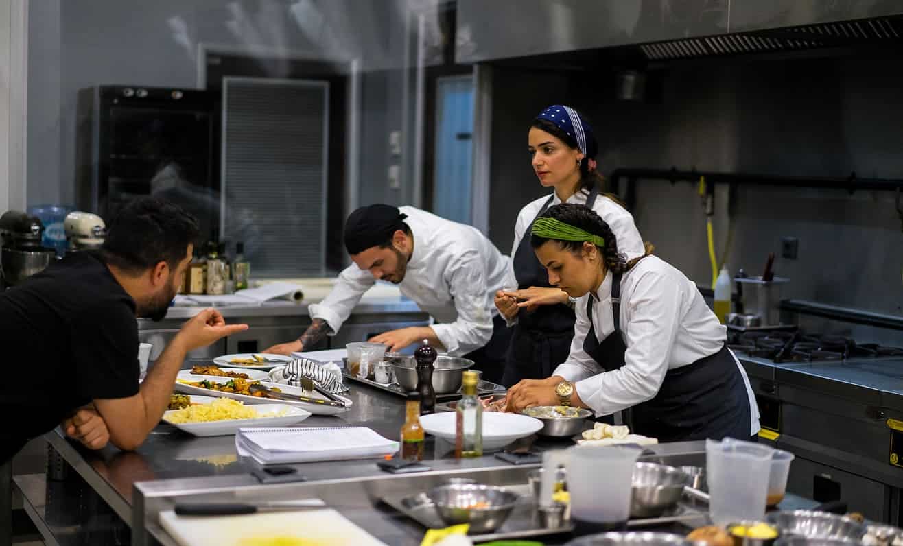 Equipe de cozinheiros, composta por homens e mulheres, cozinham em uma cozinha industrial.