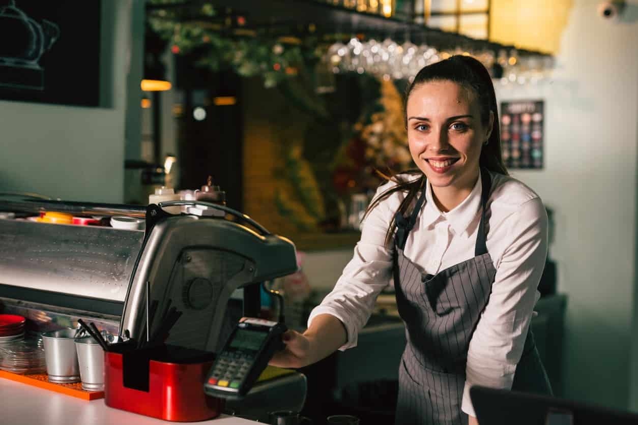 Uma moça jovem sorrindo para foto enquanto usa um avental de uniforme do restaurante e está no balcão do caixa.
