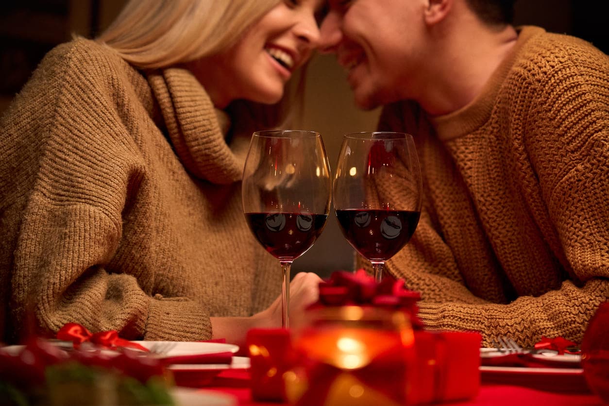 Casal em clima romântico ao fundo da foto com duas taças de vinho tinto na mesa.