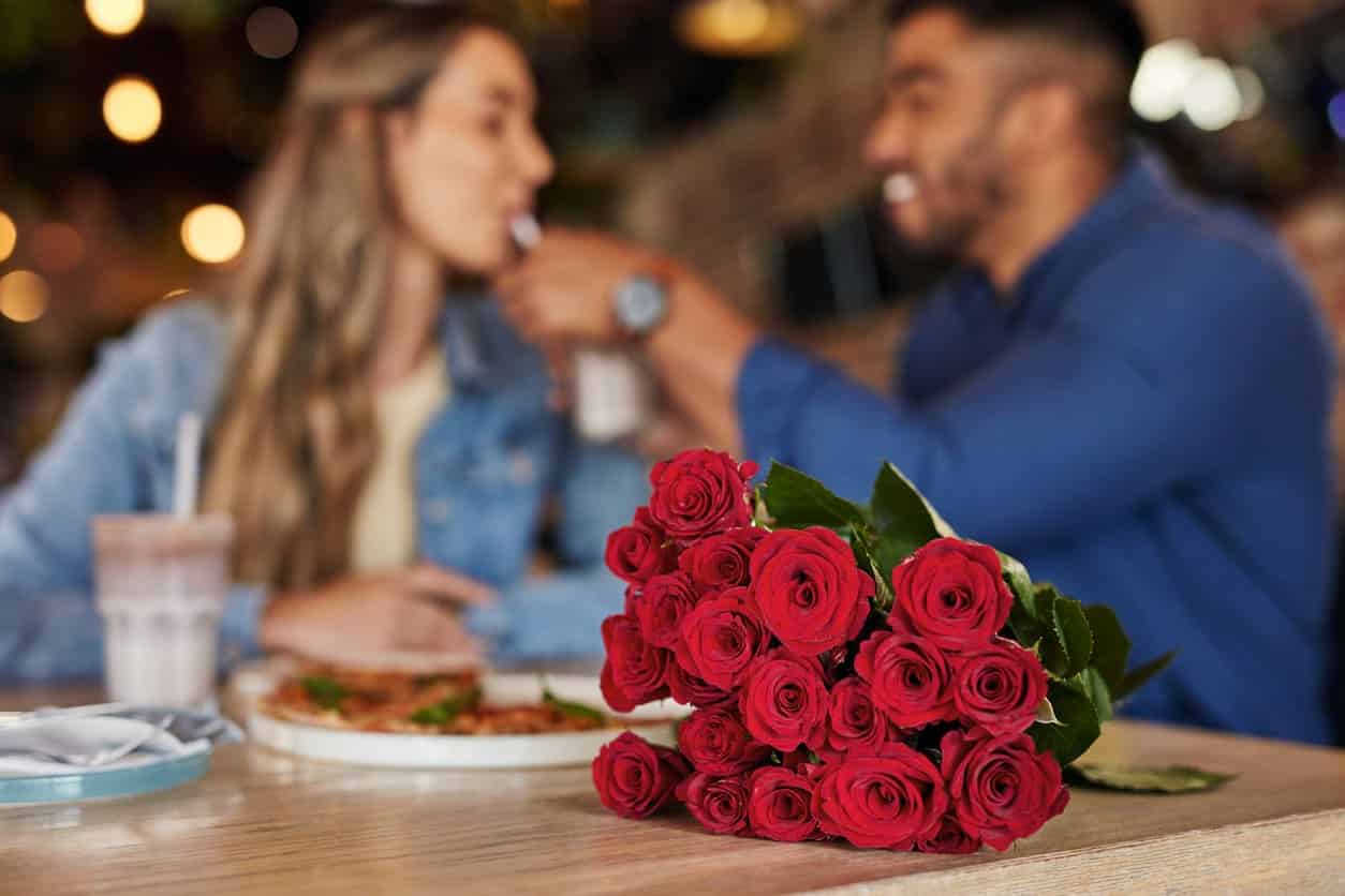 Casal em restaurante ao fundo desfocado com um buquê de rosas vermelhas na mesa.
