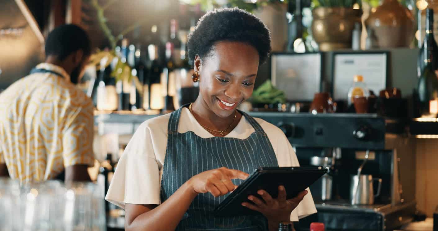 Mulher sorridente usando um tablet em um restaurante, vestindo avental azul e blusa branca, ambiente aconchegante e bem iluminado.