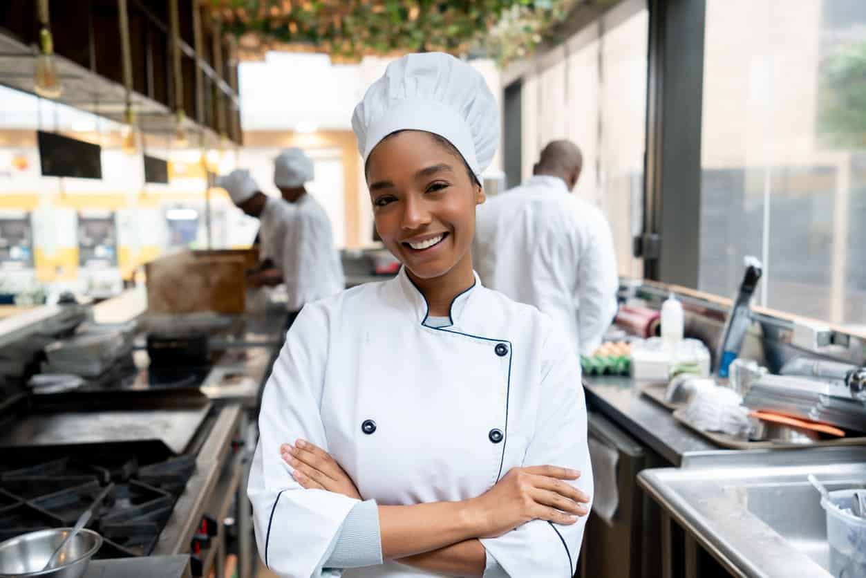 Cozinheira chefe que trabalha em uma cozinha em um restaurante posando para a foto sorrindo e com braços cruzados na altura do peito.