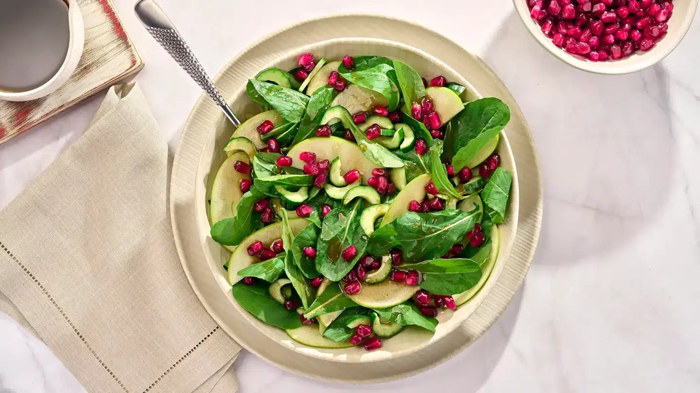 Salada com romã em um prato cor de creme disposto em uma mesa com uma toalha branca e um pote apenas com romãs ao lado.
